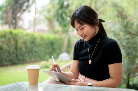 An attractive Asian woman sits at an outdoor table in a green garden or backyard using her digital tablet, working remotely. people and wireless technology conceptsの写真素材