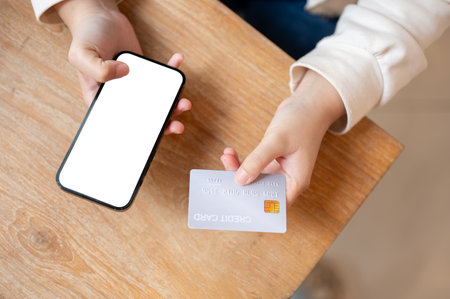 A top view image of a woman holding a credit card and a smartphone over a wooden table, a smartphone white screen mockup to display your graphic ads. online shopping, online payment, cashlessの写真素材