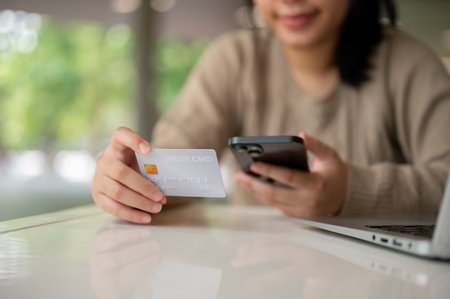 A cropped image of a young happy Asian woman holding her smartphone and a credit card, using her mobile banking app while sitting at a table indoors. online shopping, online payment, cashlessの写真素材