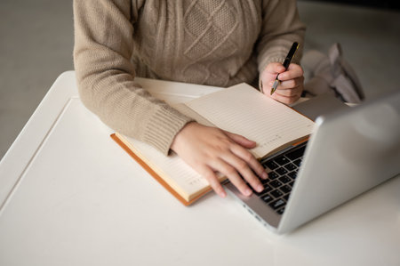 A cropped image of an Asian woman in cozy knitted sweater taking notes in her notebook while working on her laptop computer at a table indoors.の写真素材