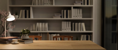 A long wooden table features books, a table lamp, a coffee cup, a potted plant, and a copy space against a large bookshelf in a reading room or office at night. 3d render, 3d illustrationの写真素材