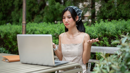 A beautiful Asian woman is having an online meeting with her colleagues, working remotely in a green garden outdoors. people and technology conceptsの写真素材