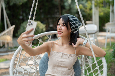 An attractive, cheerful Asian woman in a cute dress takes a selfie or records a video on her smartphone while chilling on a swing in a garden on a bright day.の写真素材