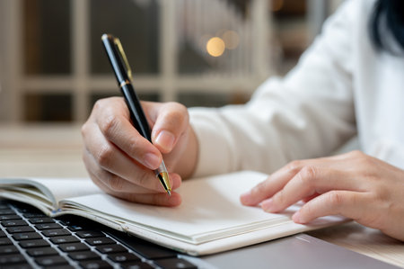 A close-up image of a woman holding a pen, writing something in her notebook while working on her laptop computer indoors. A student studying online on her laptop and taking notes in a book.の写真素材