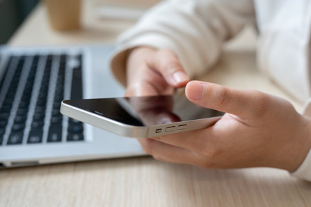A close-up image of a woman using her smartphone at her office desk, checking messages or using a...