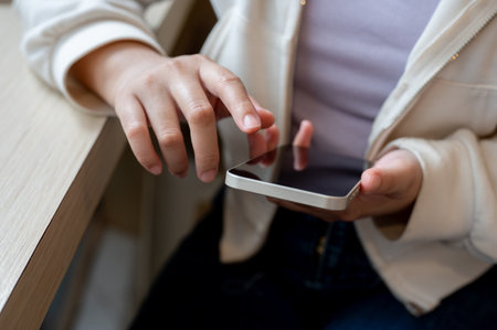 A close-up image of a woman in casual wear using her smartphone at a table indoors, reading online...