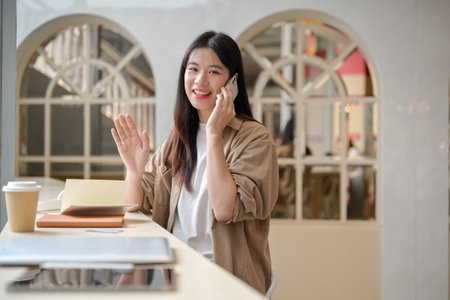 An attractive young Asian woman is waving her hand to greet someone while talking on the phone in a coffee shop, smiling at the camera. people and wireless technology conceptsの写真素材