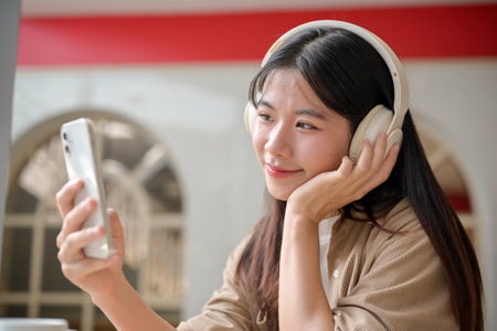 An attractive, happy young Asian woman is scrolling on her smartphone while enjoying listening to music on her headphones in a cafe. people, wireless technology, and lifestyle conceptsの写真素材