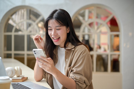 A young cheerful Asian woman is sitting in a cafe and looking at her smartphone with an excited expression. surprised by online shopping discount, reading an unexpected messageの写真素材