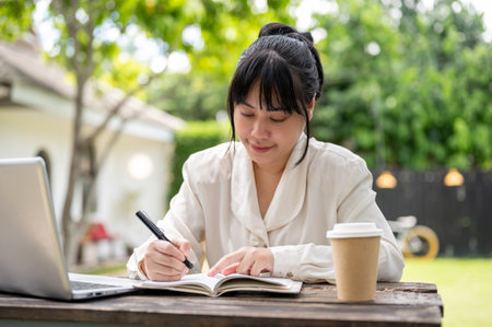 A positive Asian businesswoman is working remotely from home, sitting at a wooden table in her backyard with her laptop, focusing on her work and writing ideas in her notebook.の写真素材
