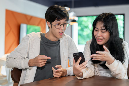 Two young Asian friends are looking at a smartphone screen and talking while sitting together in a coffee shop or a library co-working space.の写真素材