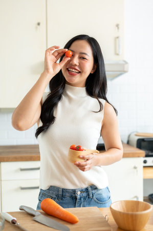 A beautiful, attractive young Asian woman enjoying cooking in the kitchen, preparing her healthy salad, posing with a tomato. home cooking, healthy lifestyle, wellness eating, weight loss recipesの写真素材