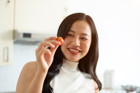 A beautiful, attractive young Asian woman enjoying cooking in the kitchen, preparing her healthy salad, posing with a tomato. home cooking, healthy lifestyle, wellness eating, weight loss recipesの写真素材