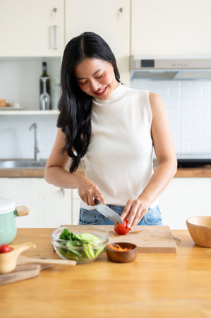 A beautiful, happy young Asian woman cutting a tomato on a cutting board, preparing ingredients for her healthy salad, enjoying cooking in the kitchen. home cooking, domestic life, healthy eatingの写真素材