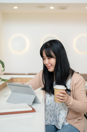 An attractive young Asian woman having a hot drink and using her digital tablet at a table in a minimalist cafe. people, wireless technology, lifestylesの写真素材