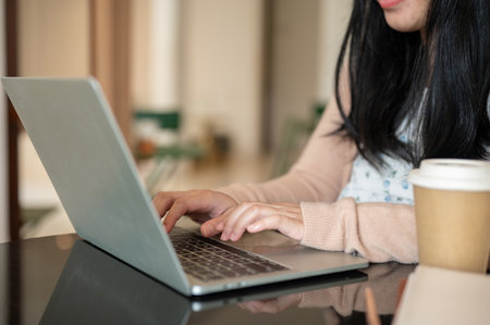 A cropped image of a woman working on her laptop computer at a table indoors, typing on the laptop...