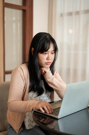 A serious, thoughtful Asian woman is focusing on working on her laptop computer, reading an online...