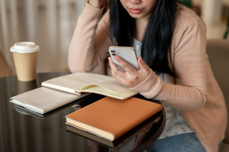 A cropped image of an Asian woman using her smartphone at a table indoor while reading books. people and technology conceptsの写真素材