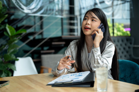 A confident, successful Asian businesswoman or female boss is talking on the phone with her client while reviewing document at her desk in the company building.の写真素材