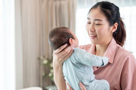 A happy young Asian mom is talking and comforting her infant baby son while holding him in her arms, standing by the window in the living room. mother's love and tenderness, motherhoodの写真素材