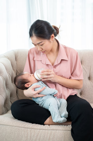 A caring young Asian mom is holding her newborn baby son in her arm and feeding him milk from a bottle on the sofa in the living room. motherhood and childcare conceptsの写真素材