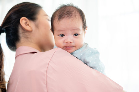 An Asian mom is cuddling her newborn baby while standing by the window indoors, having a great time...