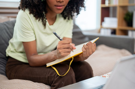 A cropped image of a Black woman writing her diary or taking notes while studying online on her laptop, on a couch in the living room.の写真素材