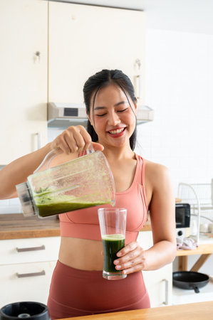 An attractive, healthy Asian woman in sportswear is pouring her green juice from a blender, making her healthy drink after workout at home. healthy lifestyle, domestic life, wellness livingの写真素材
