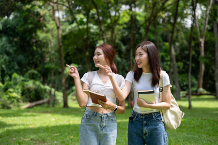 Two pretty and happy young Asian female college students with backpacks are pointing their finger at something, talking, and walking in the green park together.の写真素材