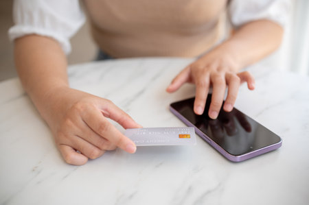 A close-up image of a woman sitting at a table indoor with her smartphone and credit card, shopping...