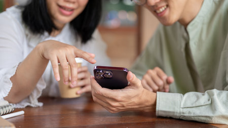 A close-up image of two happy Asian friends are enjoying talking while watching a video on a smartphone together, relaxing at a coffee shop in the city. people, lifestyle, and wireless technologyの写真素材