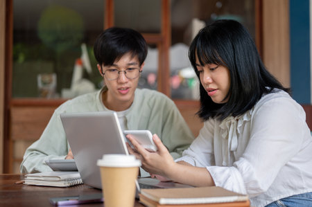 A young Asian woman is using a calculator while discussing work with her male colleague at a coffee shop, working remotely, having an informal meeting, brainstorming.の写真素材