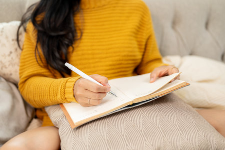 A close-up image of an Asian woman in a cozy yellow knitted sweater writing her diary on a couch at...