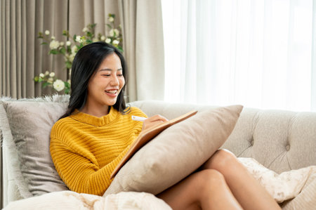 A young, positive Asian woman in comfy clothes is enjoying writing in her diary on a couch at home. leisure, lifestyle, hobby, domestic lifeの写真素材