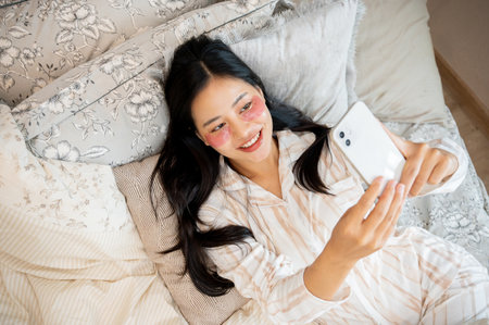 A top view image of a beautiful, happy Asian woman in pajamas and with eye patches on her face lying in bed and using her smartphone. people and domestic life conceptsの写真素材