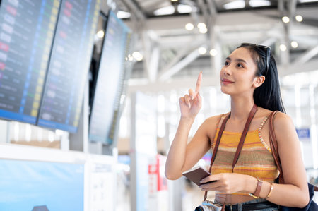 A charming young Asian female tourist is checking her flight boarding time at a flight information display in the airport terminal. traveling, lifestyle, transportationの写真素材