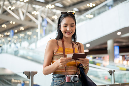 A charming, smiling Asian female tourist passenger is looking at her passport in her hand, standing in the airport terminal, traveling by plane. people and transportation conceptsの写真素材