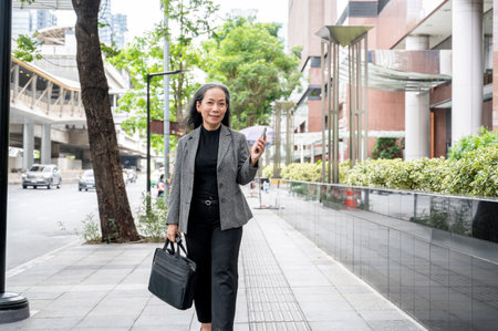 A confident, positive mature Asian businesswoman is walking on a sidewalk in the city, holding her smartphone and a briefcase, commuting to work in the morning.の写真素材