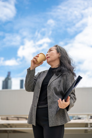 A confident, successful Asian senior businesswoman in a formal business suit stands in the urban city surrounded by skyscraper buildings, sipping a coffee.の写真素材