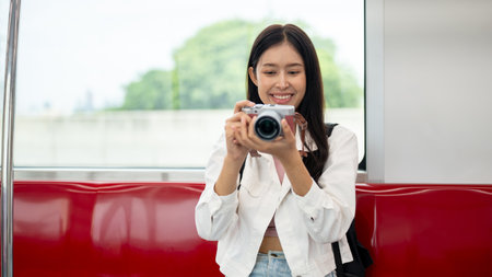 A cheerful, charming young Asian female tourist is taking pictures with her camera while commuting in a city on a sky train, enjoying her time on public transport.の写真素材