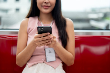 A cropped image of a happy Asian businesswoman or female office worker sitting on a seat and chatting on her smartphone while taking the sky train to work, using public transport in an urban city.の写真素材