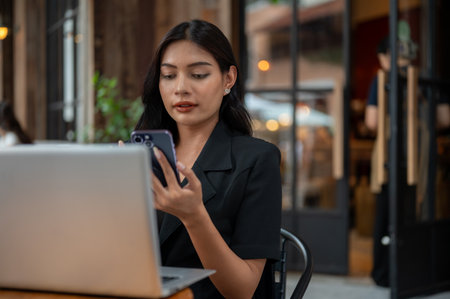 An attractive Asian businesswoman in a black suit is using her smartphone, reading messages, while working on her laptop and working remotely from a cafe in the city.の写真素材