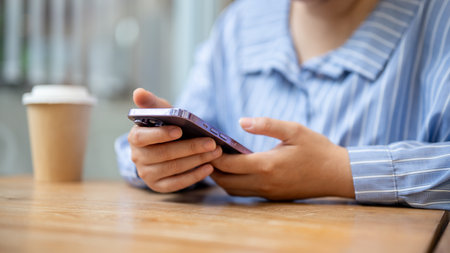A cropped image of an Asian woman using her smartphone while sitting at a table in a coffee shop. using a mobile app, chatting, reading online news, scrolling on social mediaの写真素材