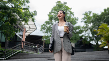 A beautiful, confident Asian businesswoman in a stylish suit is walking down the stairs in the city with a takeaway coffee cup in her hand, going to the office in the morning.の写真素材