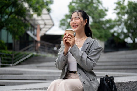 A beautiful, happy Asian businesswoman is having her morning coffee while sitting on the stairs in the city or relaxing on her coffee break outdoors. city life and business people conceptsの写真素材