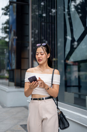 A beautiful, smiling Asian woman in a trendy outfit is standing in front of the shopping mall with a...