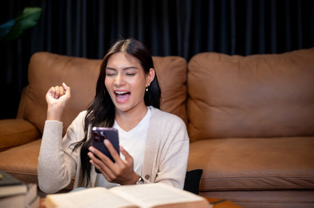 A cheerful, excited Asian woman in comfy clothes is screaming with joy at her smartphone and showing her victory fist, celebrating good unexpected news, sitting in the living room at night.の写真素材