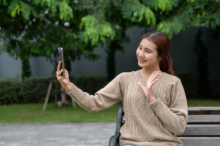 A positive, charming young Asian woman is taking a selfie or talking on a video call with her friend while resting on a bench in a park. people and wireless technology conceptsの写真素材