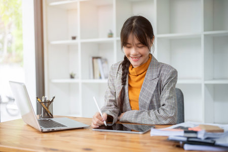 A charming, positive Asian businesswoman or female office worker is using a digital tablet at her...