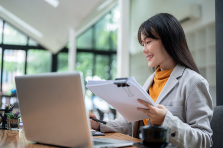 A satisfied, beautiful young Asian businesswoman is working in her office, reviewing business reports and managing business documents at her desk.の写真素材